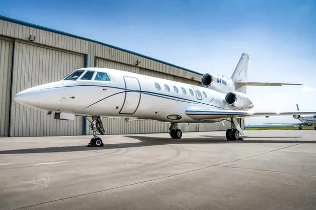 A front-side view of a white Dassault Falcon 900EX business jet with blue stripes, registration N47NS, parked on an apron.