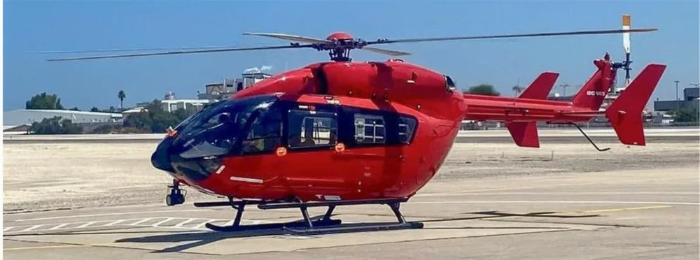 A red Airbus H145 helicopter is parked on a tarmac with markings under a clear sky.
