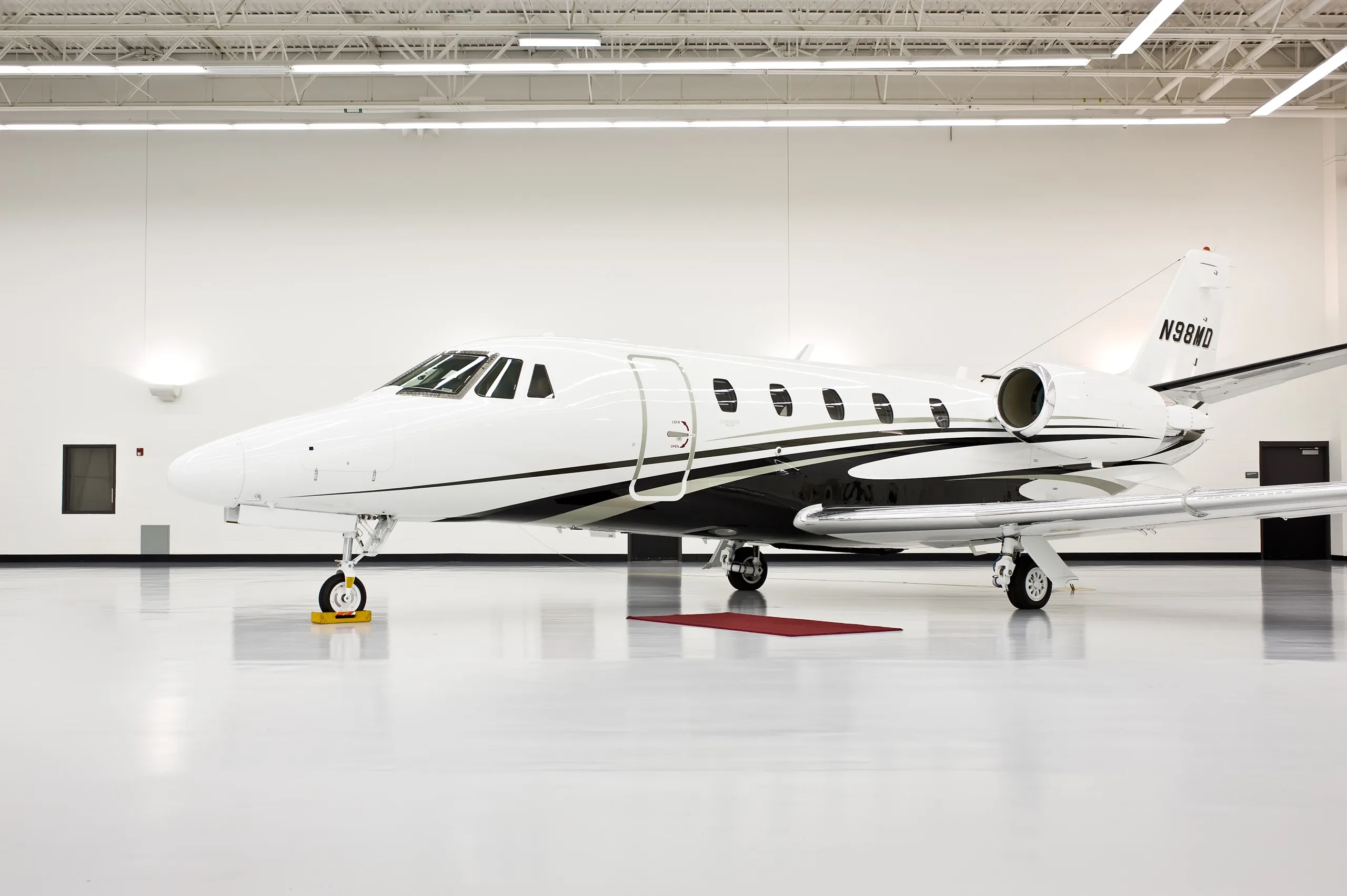 A white business jet with black and grey stripes, registration N98MD, parked in a brightly lit hangar.