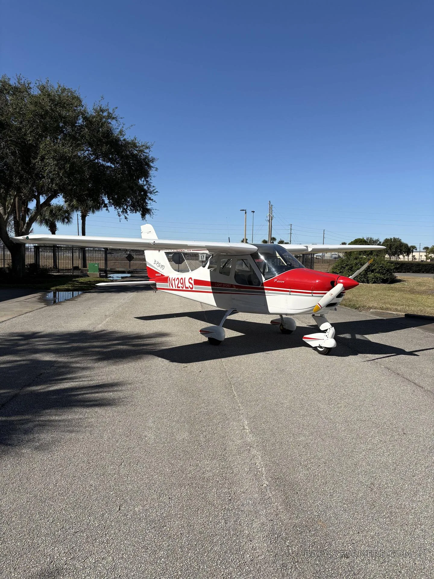 A white and red Tecnam P2008 aircraft with registration N129LS is parked on an asphalt surface next to a tree under a clear blue sky.