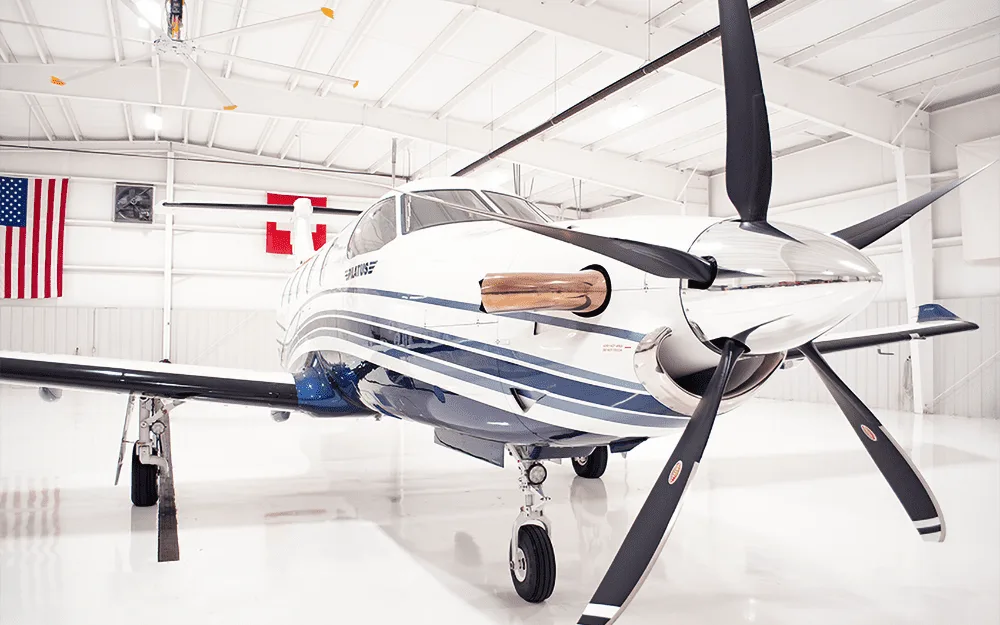A close-up front view of the propeller and nose of a white and blue Pilatus PC-12 aircraft in a hangar.