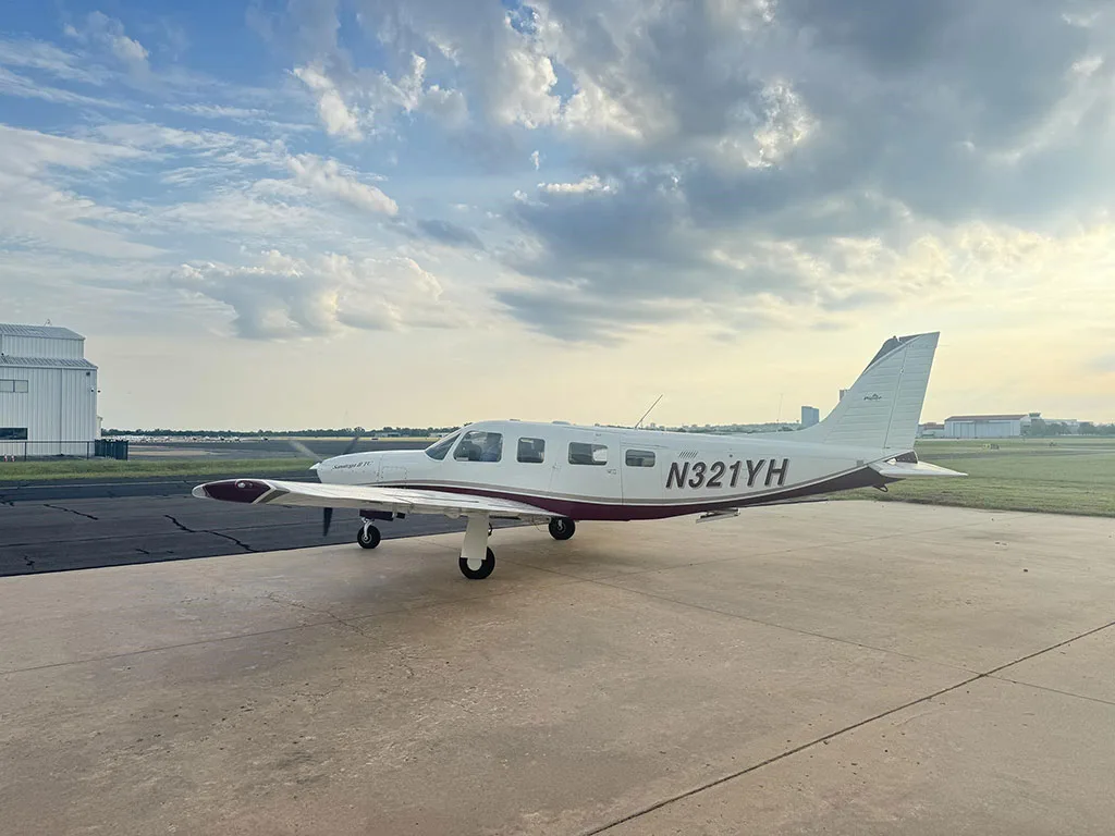 A white and maroon Piper Saratoga II TC aircraft (N321YH) parked on an airport apron with distant buildings under a partly cloudy sky.