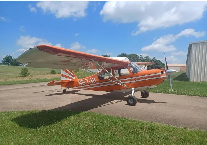 A bright orange single-engine aircraft, N57411, with white stripes and star decals, parked on an asphalt ramp under a blue sky.