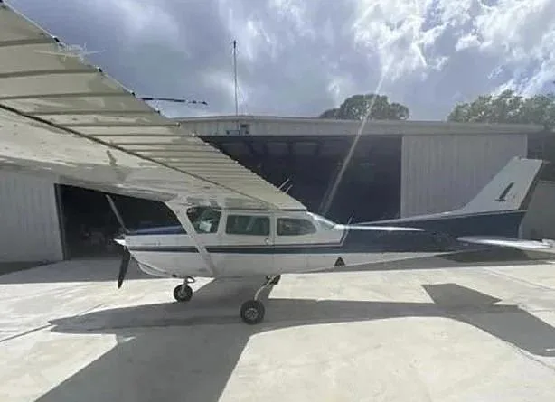 A white and blue single-engine propeller aircraft (Cessna) parked on a tarmac in front of a hangar under a cloudy sky.