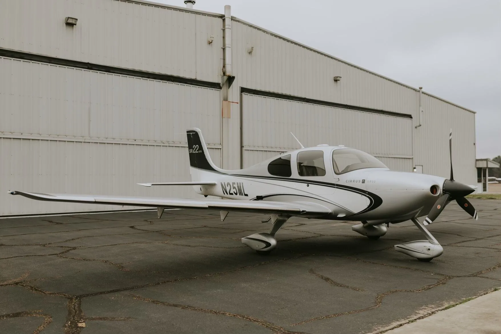 A silver Cirrus SR22 GTS aircraft with black and white stripes parked on an asphalt ramp in front of a hangar.