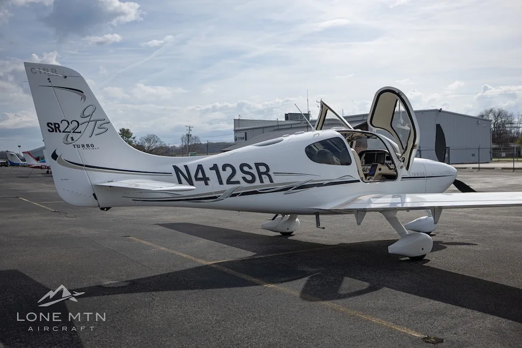A white Cirrus SR22T aircraft with open doors is parked on an asphalt runway next to airport buildings.