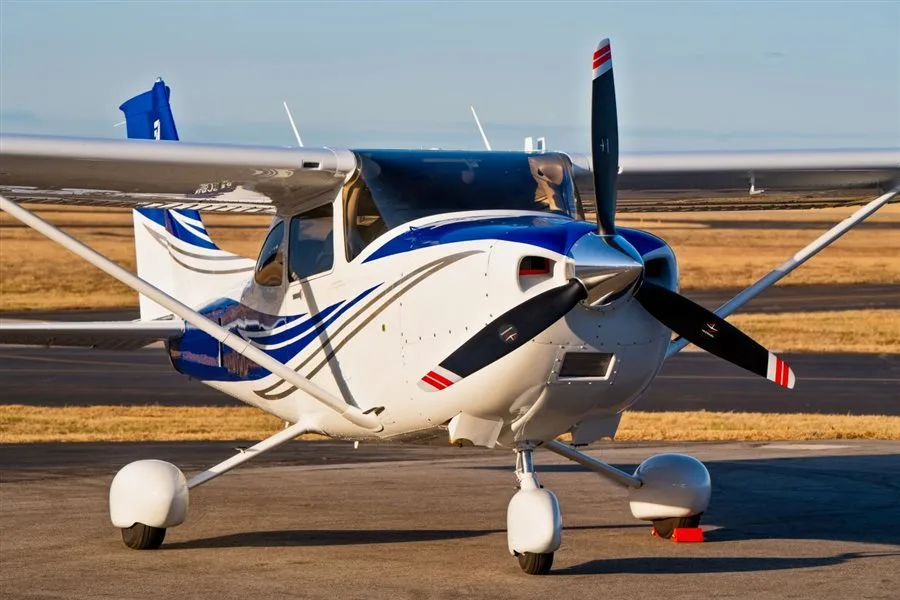 A white single-engine propeller aircraft with blue and silver stripes, parked on an airfield with dry grass in the background.