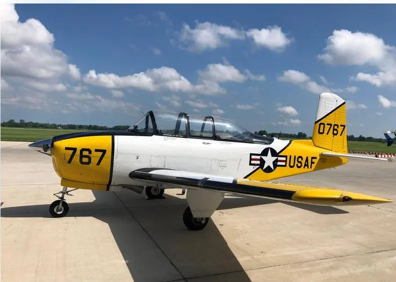 A yellow and white military trainer aircraft with USAF markings and number 767, parked on an airfield under a partly cloudy sky.