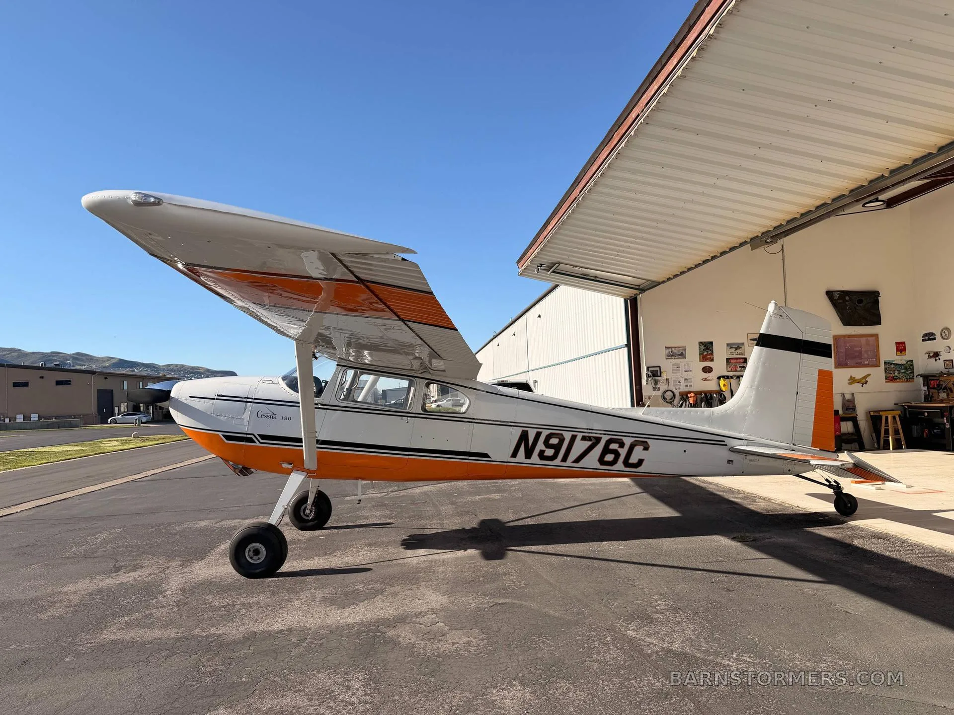 A white and orange Cessna 180 aircraft, registration N9176C, parked on an asphalt ramp with a hangar and hills in the background under a clear blue sky.