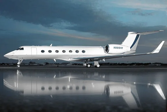 A white Gulfstream G650 business jet with a blue tail stripe is parked on a reflective tarmac under a dramatic sky.