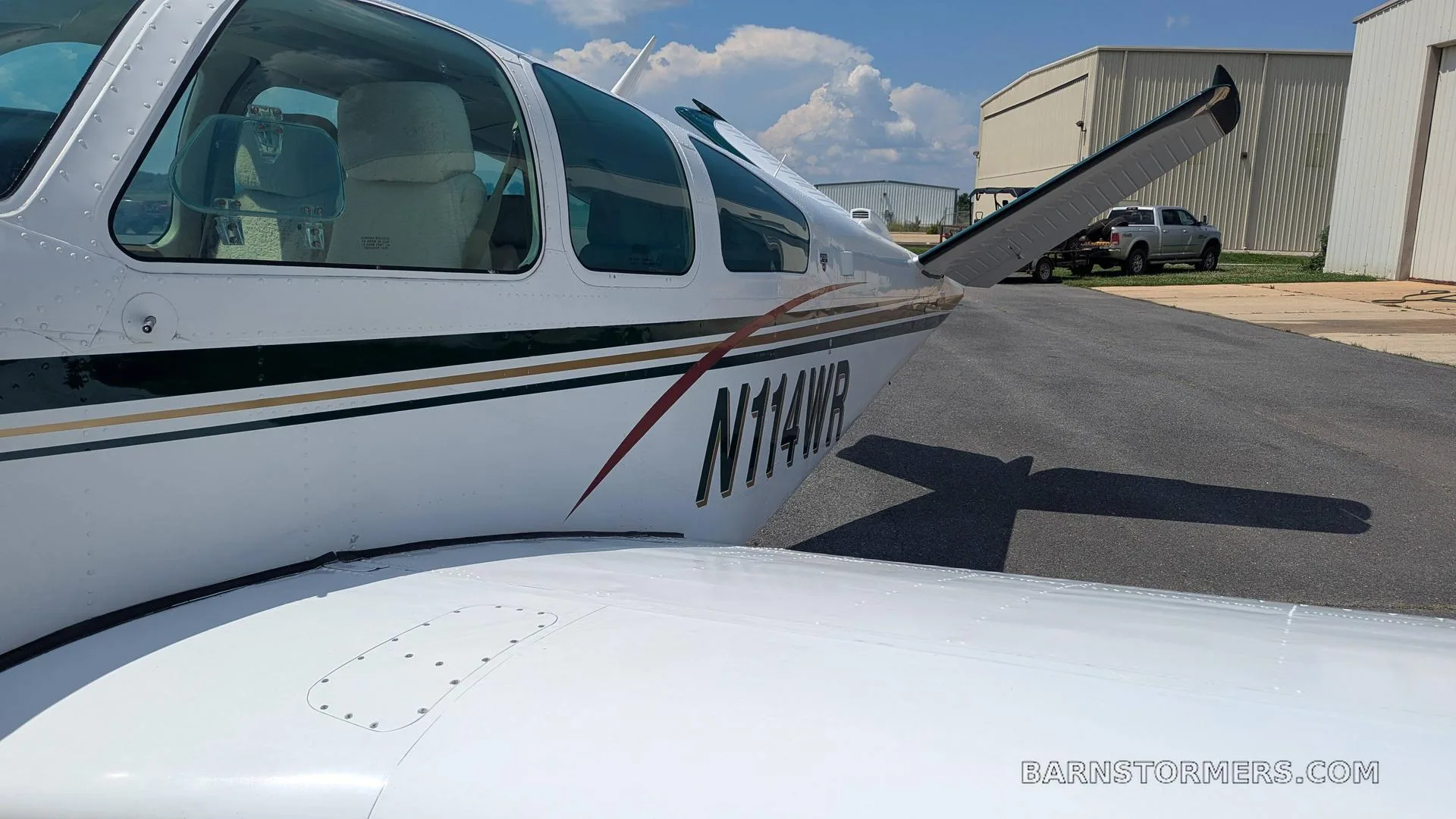 A close-up side view of the white Beechcraft Bonanza's fuselage and tail, showing the N114WR registration and green/gold stripes.
