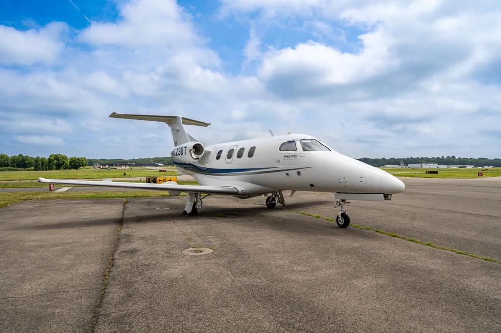 A white Embraer Phenom 100 business jet with blue and grey stripes, registration N623DT, parked on an airfield under a partly cloudy sky.