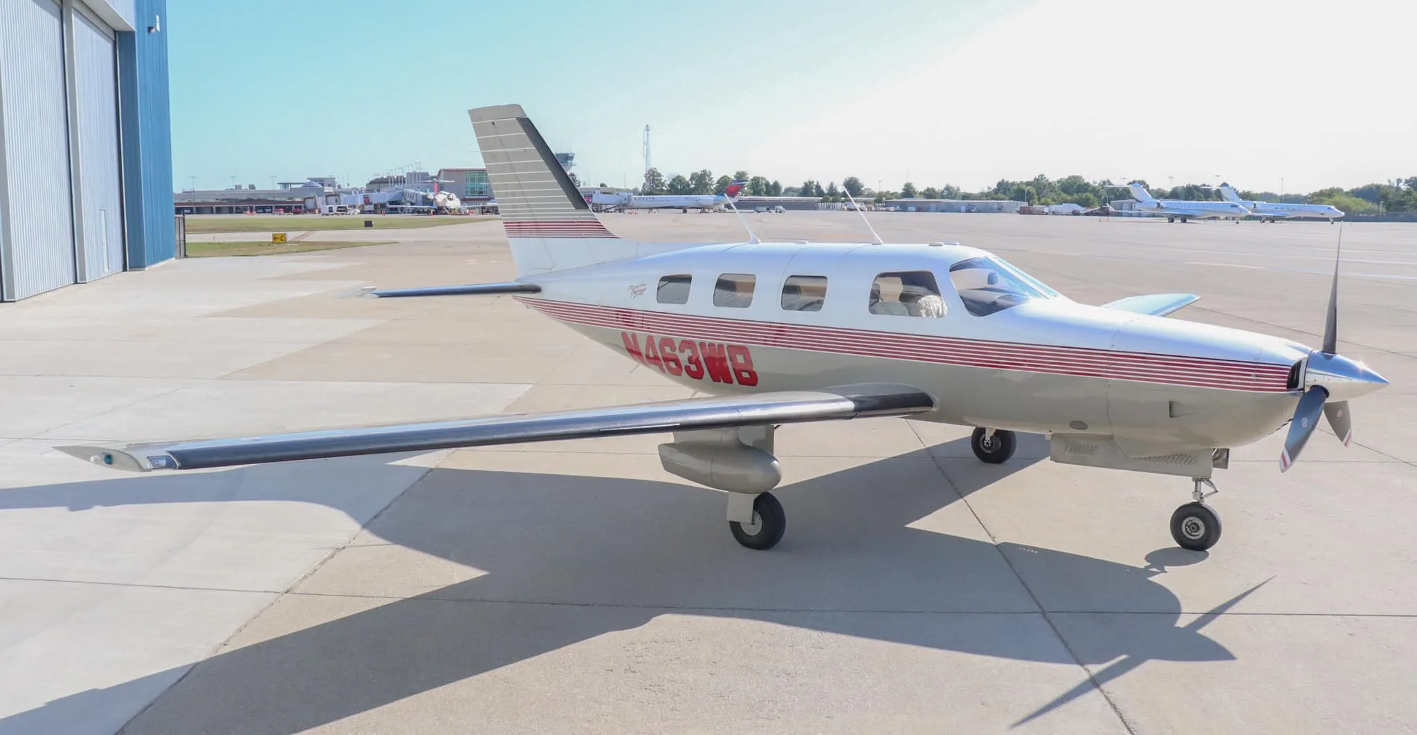 A white and tan Piper Malibu Mirage N463WB with red stripes is parked on an airport tarmac with other aircraft in the background.