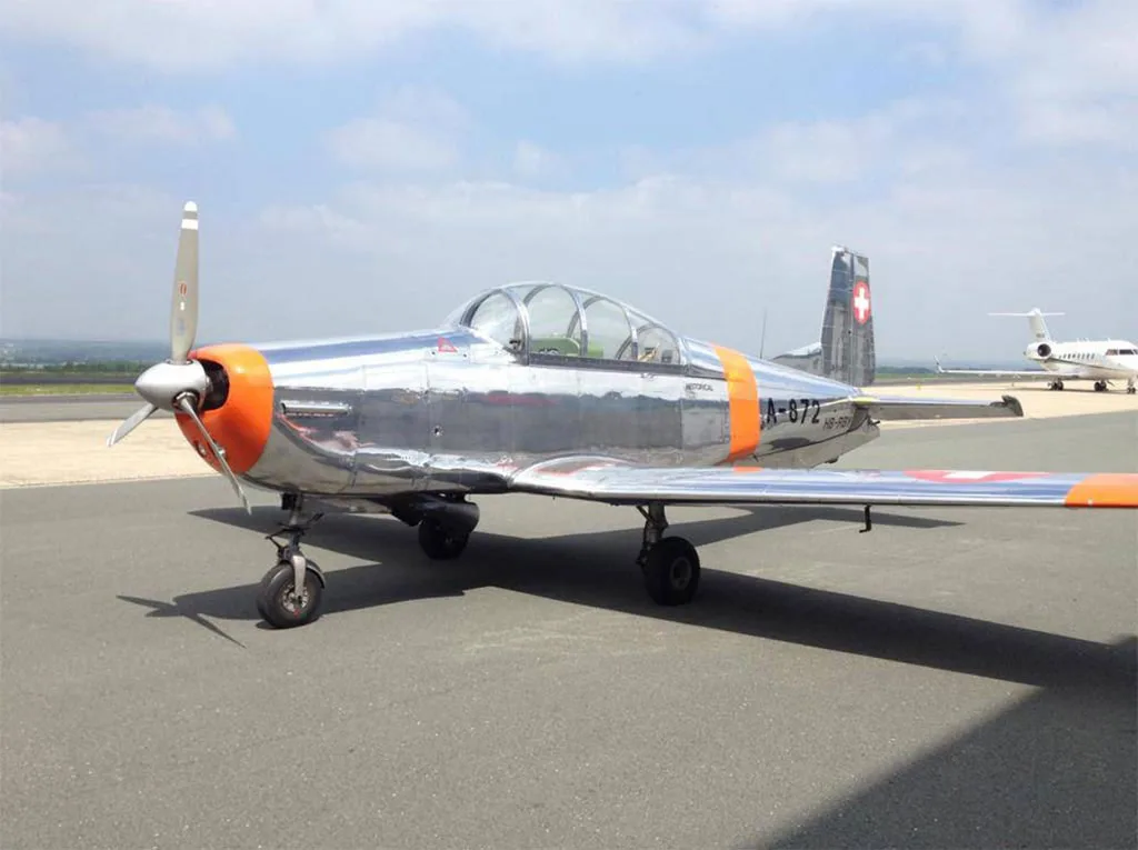 A polished silver and orange Pilatus P-3 aircraft with Swiss markings, parked on an airfield under a partly cloudy sky.