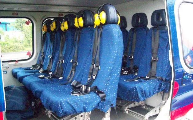 A view of the helicopter's interior cabin shows rows of blue seats with yellow headsets and safety harnesses.