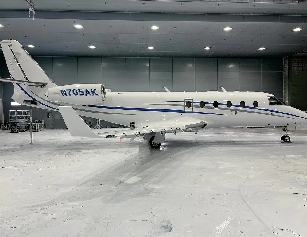 A white business jet, registration N705AK, with blue and grey stripes, parked in a hangar.