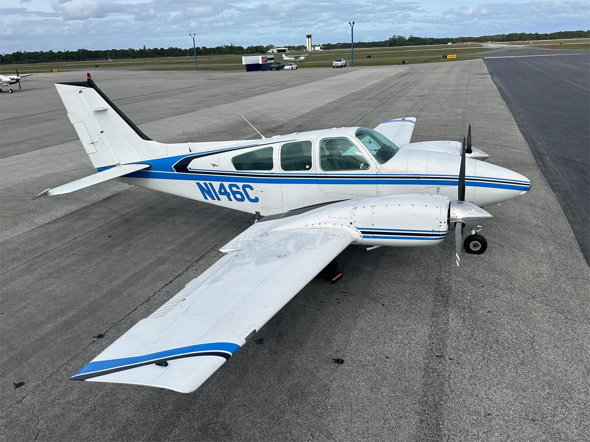 A white and blue twin-engine propeller aircraft, likely a Beechcraft Baron with registration N146C, parked on an airport tarmac under a cloudy sky.