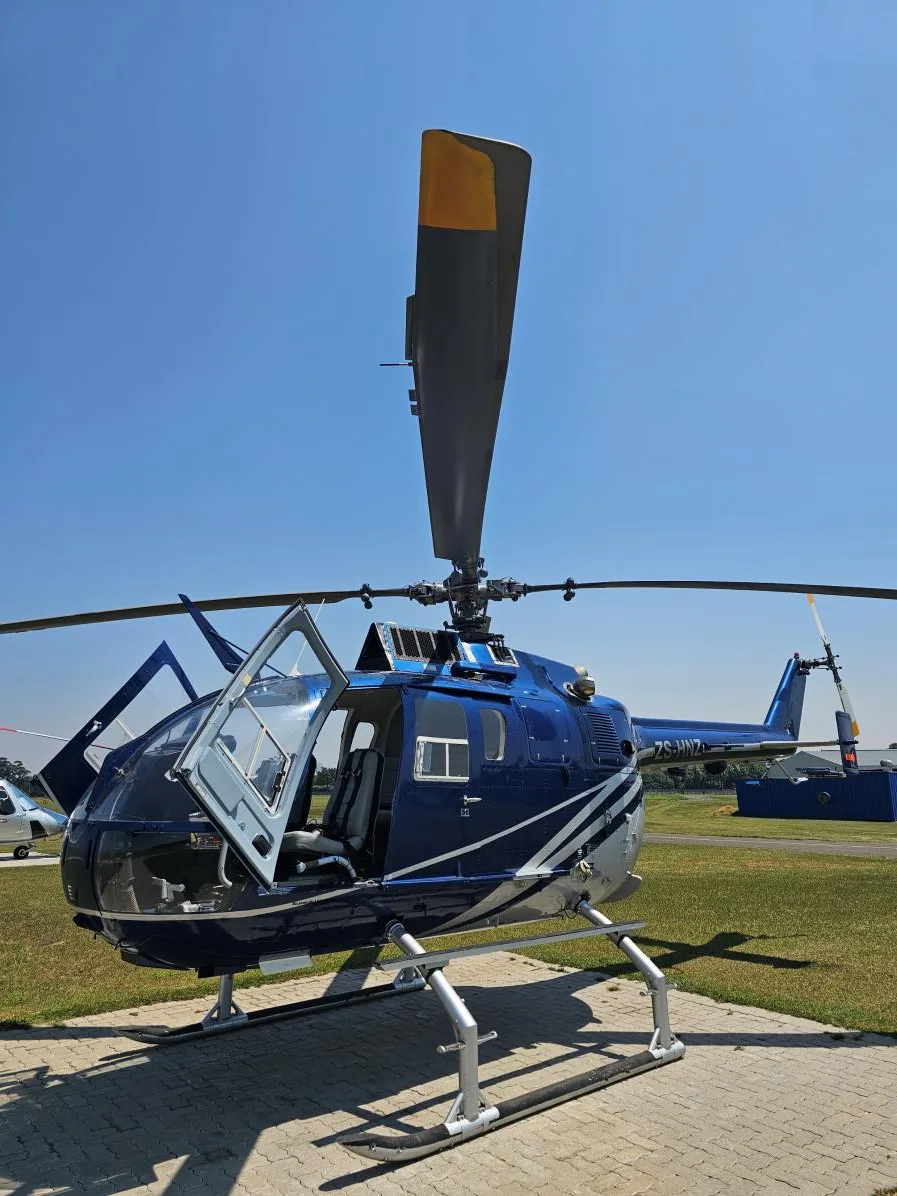 A blue and silver helicopter with open doors is parked on a paved surface at an airfield under a clear sky.