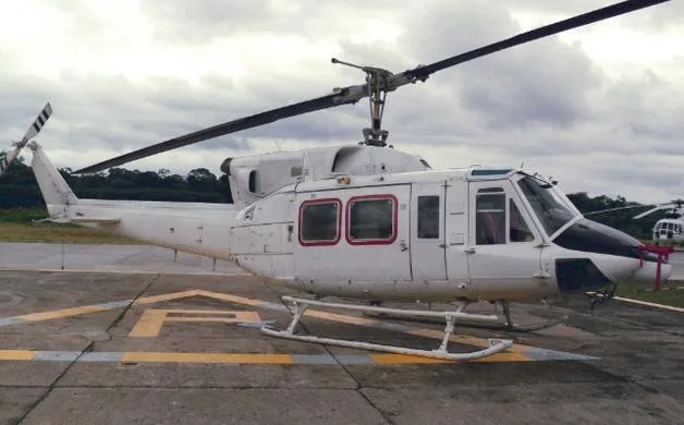 A white Bell 212 helicopter with a black nose and red window frames sits on a concrete pad with yellow markings under a cloudy sky.