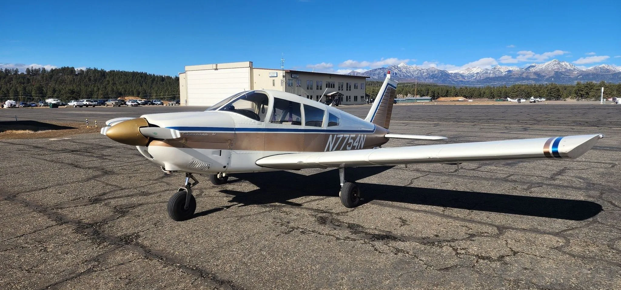 A front-left view of a white and gold Piper Cherokee 140 (N7754N) on an airfield with a building and mountains in the background.
