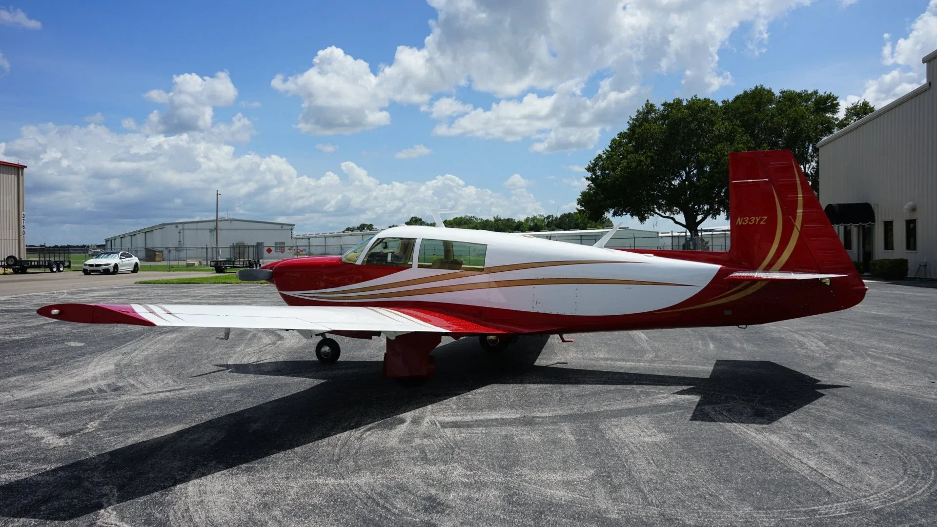 A side view of a red and white single-engine aircraft with gold stripes parked on an asphalt apron under a blue sky.