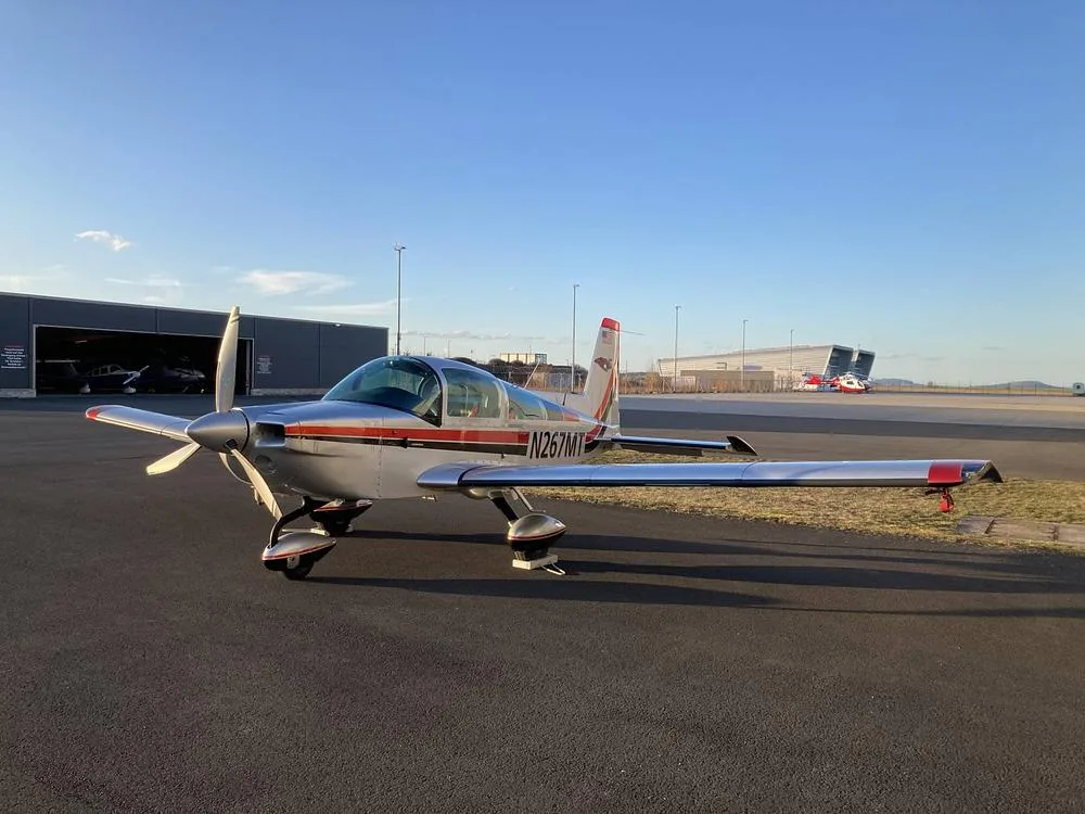 The Grumman American AA-5B Tiger aircraft, N267MT, parked on an airfield with hangars in the background under a clear sky.