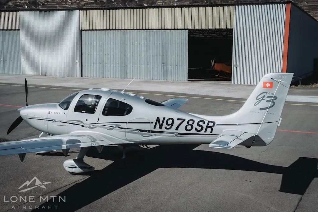 A silver Cirrus SR22 aircraft (N978SR) parked on an airfield with a hangar in the background.