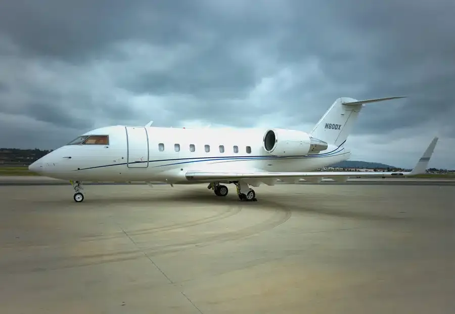 A white Bombardier Challenger business jet with blue accent stripes is parked on an airport tarmac under a cloudy sky.