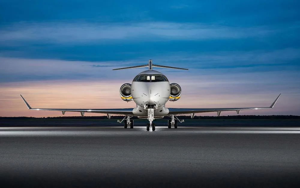 A white business jet with yellow engine accents is viewed head-on on an airfield at dusk.