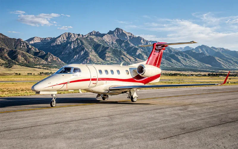 A white Learjet 75 business jet with red stripes on the fuselage and tail, parked on a runway with mountains in the background.