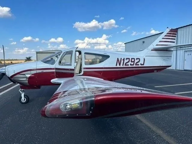 A white and red single-engine aircraft, N1292J, with its door open, parked on an asphalt apron next to a hangar.