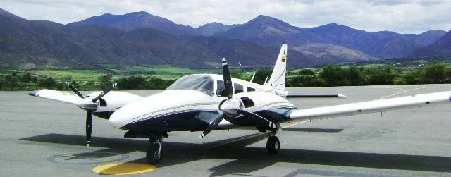 A white and blue twin-engine propeller aircraft parked on an tarmac with mountains in the background.