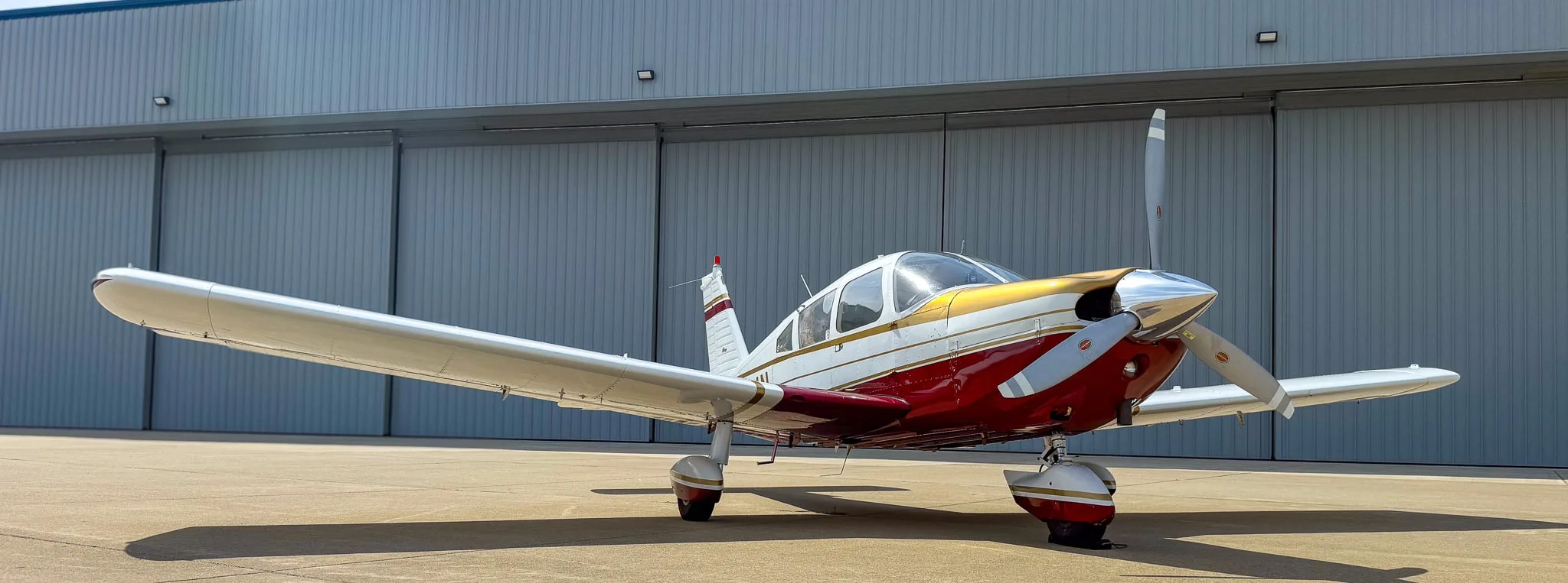 A white, red, and gold Piper Cherokee aircraft (N8534N) parked on an airfield with a hangar in the background.