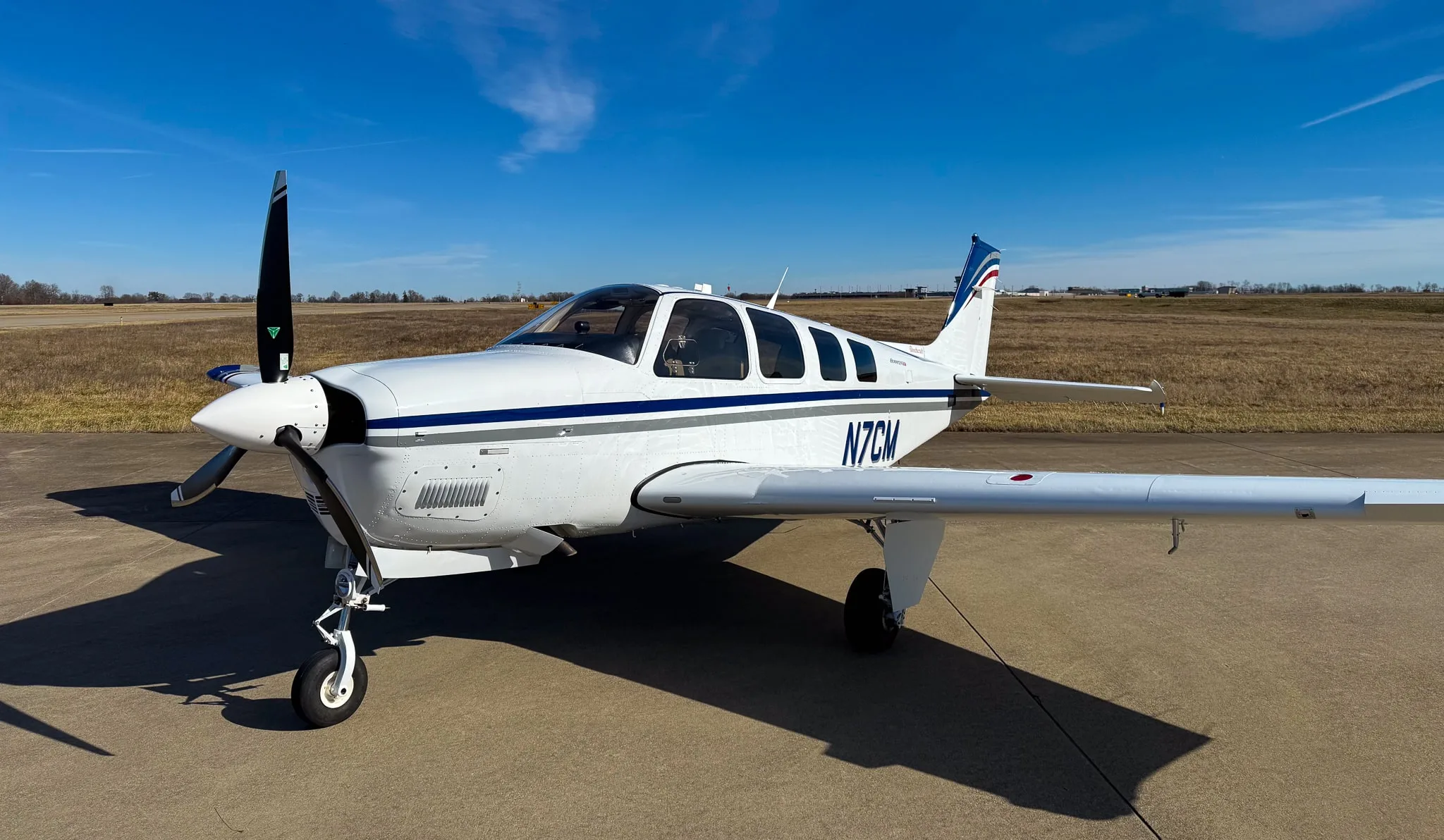 A white Beechcraft Bonanza G36 with blue and grey stripes, registration N7CM, parked on an airfield under a clear blue sky.