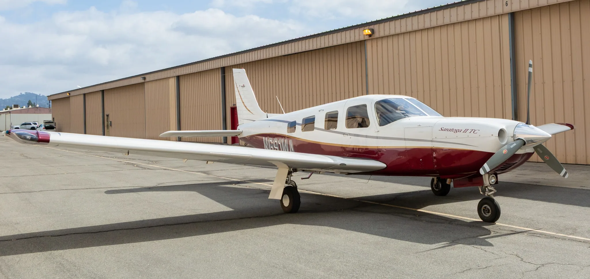 A white and maroon Piper Saratoga II TC aircraft, registration N691MA, parked on an asphalt ramp in front of hangars.