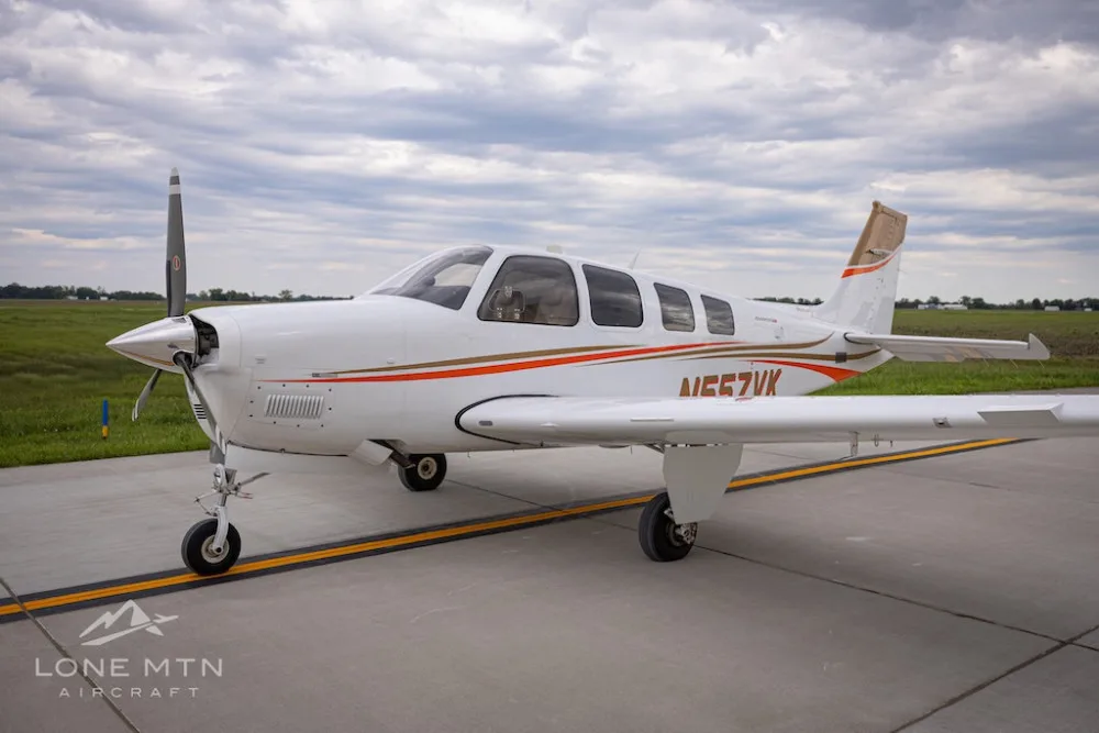 A white Beechcraft Bonanza G36 with orange and brown stripes, registration N557VK, parked on an airfield under a cloudy sky.