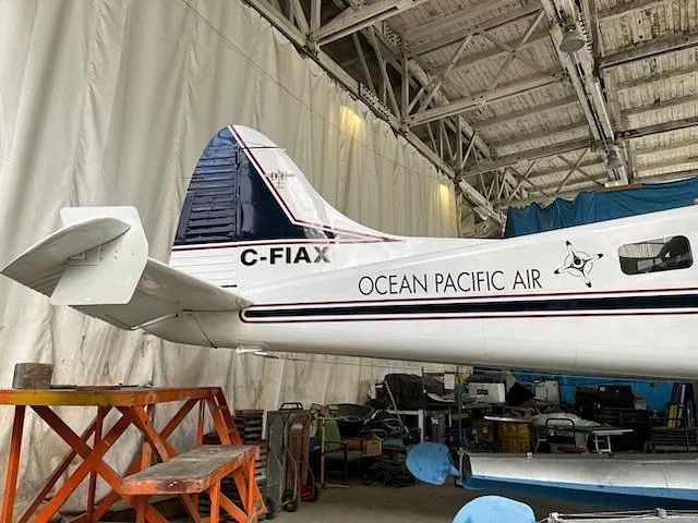 The tail section of a white De Havilland Beaver seaplane with "C-FIAX" and "OCEAN PACIFIC AIR" markings is visible inside a hangar.