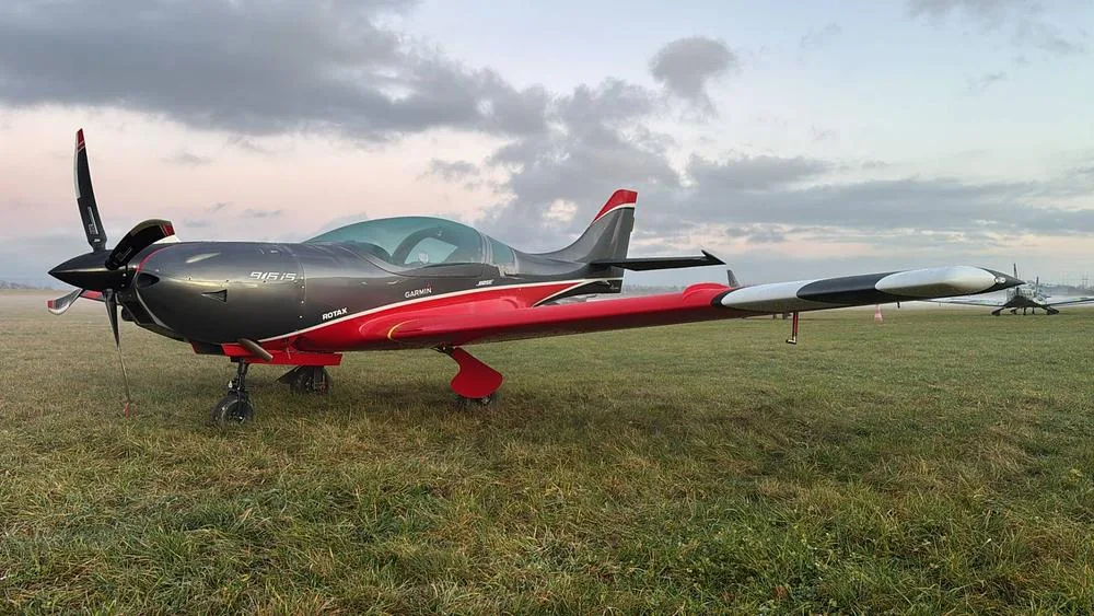A grey and red BOSSA 915iS aircraft with a propeller, parked on a grassy field under a cloudy sky.