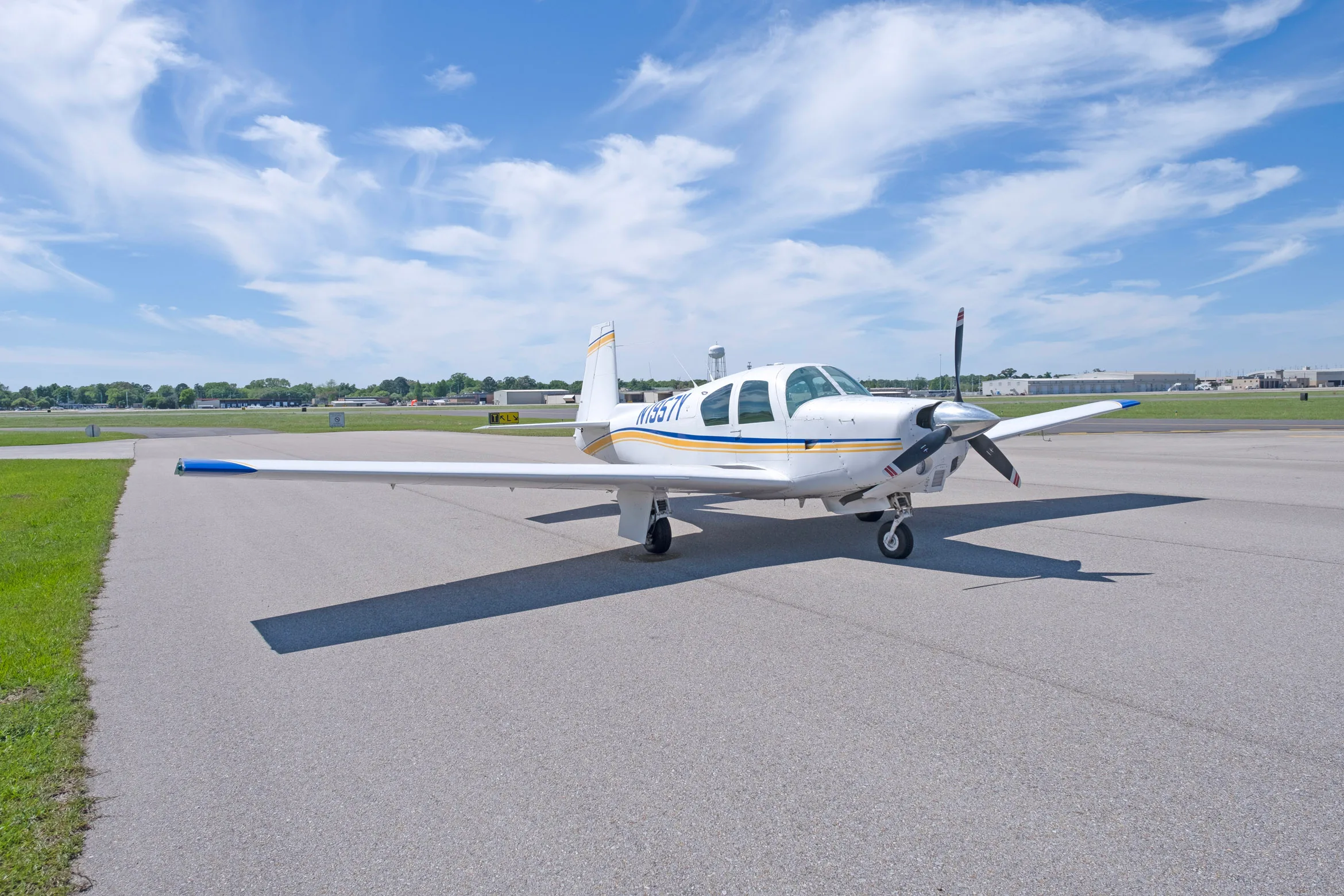 A white single-engine propeller aircraft with blue and yellow stripes, registration N1957Y, parked on an asphalt runway under a blue sky.