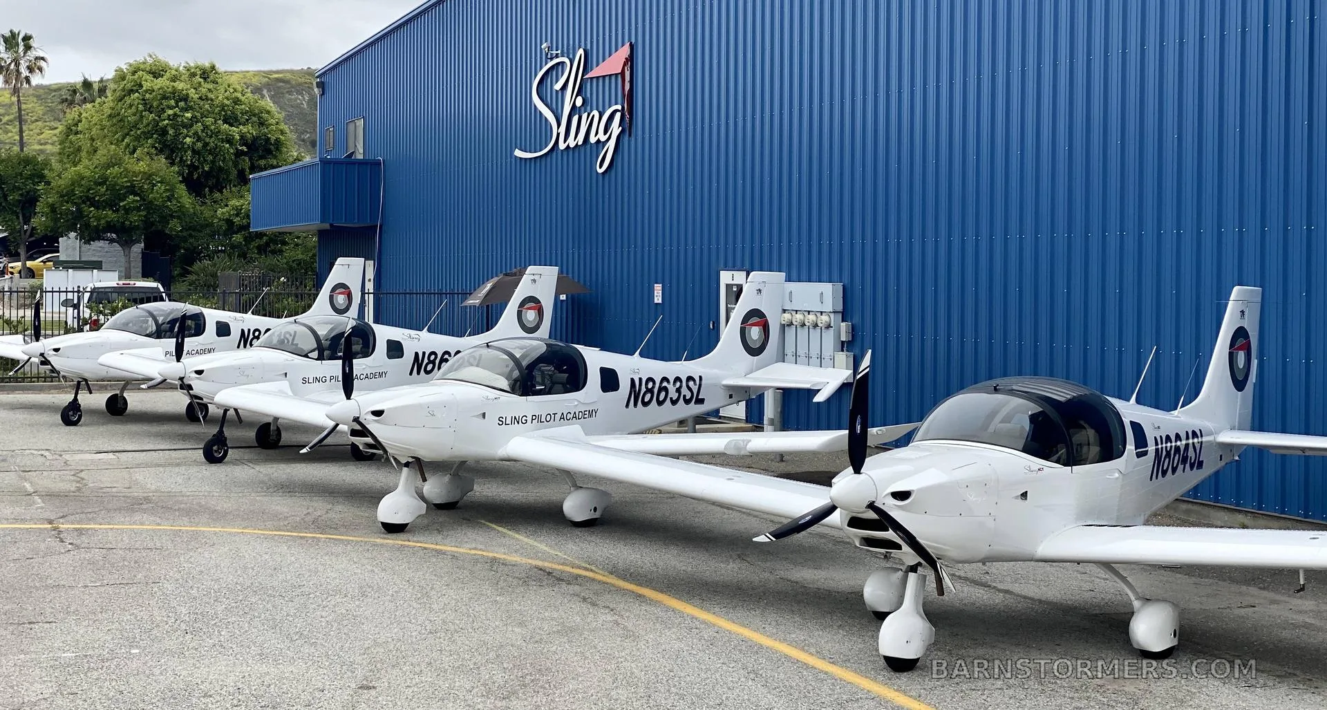 Several white Sling Pilot Academy aircraft are parked in a line in front of a blue hangar with 'The Sling' logo.