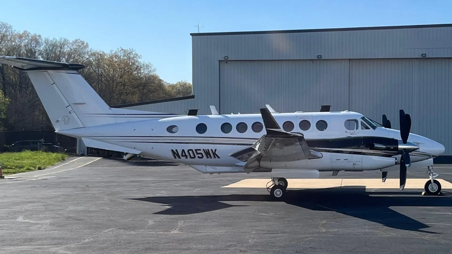 A side view of a white and black Beechcraft King Air N405NK parked on an asphalt tarmac next to a white hangar.
