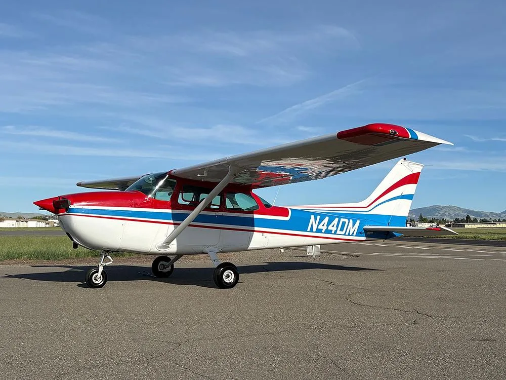 A white Cessna 172 with red and blue stripes, registration N44DM, parked on an asphalt ramp under a blue sky.