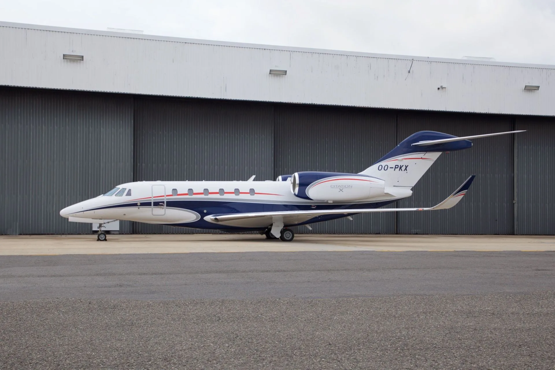 A white Cessna Citation X business jet with blue and red stripes is parked on an tarmac in front of a hangar.