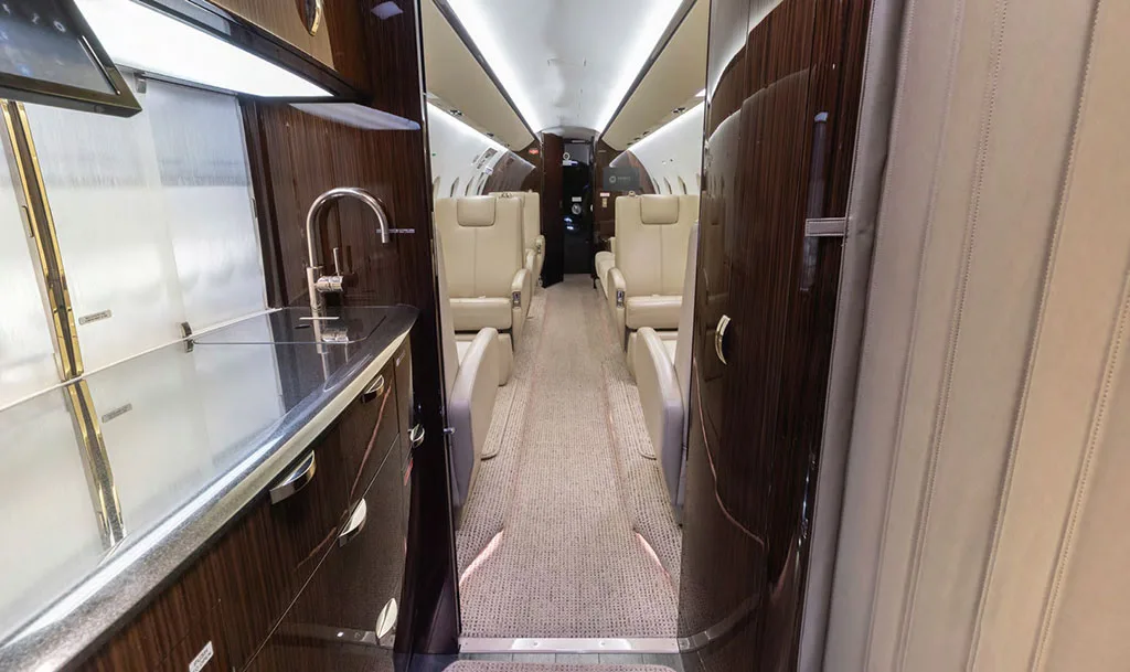 A view from the galley area into the elegant cabin of the Gulfstream G550, showing light-colored seating and dark wood cabinetry.
