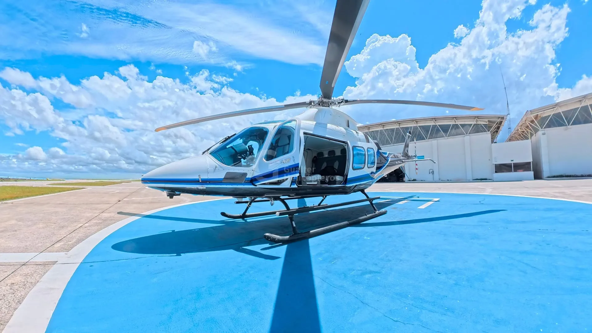 A white and blue helicopter with its door open sits on a blue helipad under a bright sky.