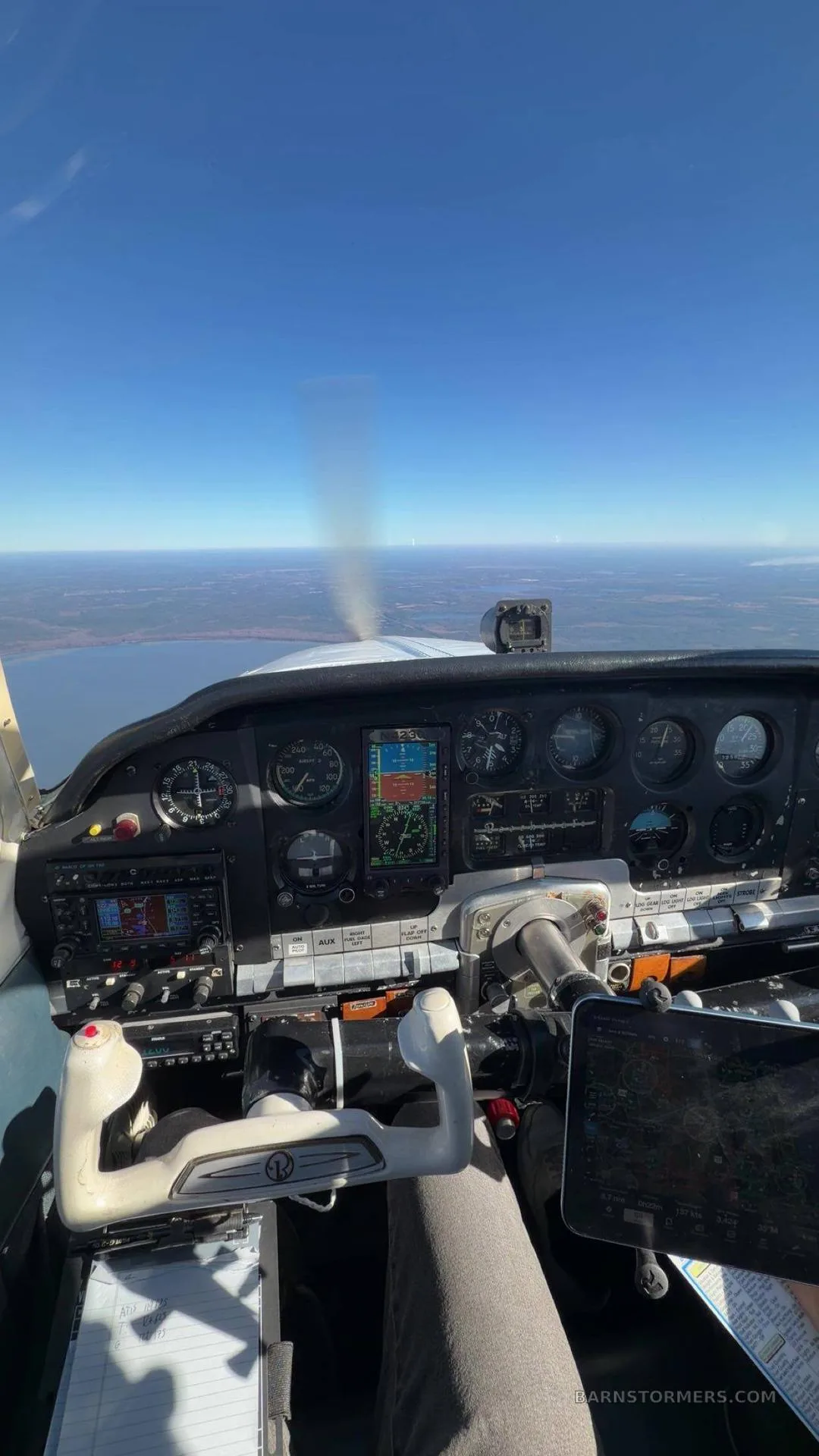 The cockpit of a small aircraft in flight, showing the instrument panel, controls, and a view of land and water below.