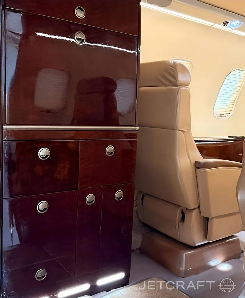 A polished dark wood cabinet with silver handles stands next to a tan leather seat in an aircraft interior.