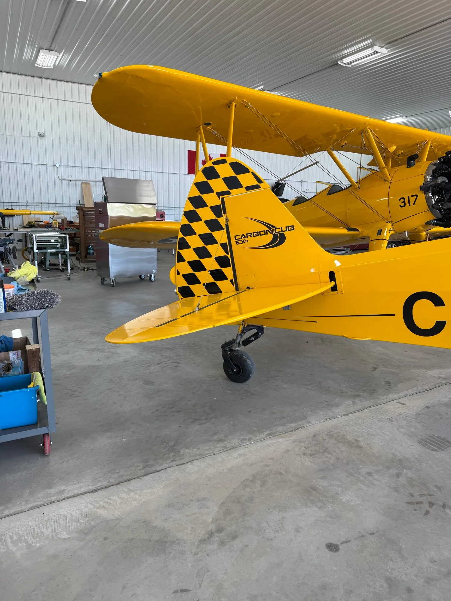 The tail section of a yellow CubCrafters Carbon Cub EX-3 with a black and yellow checkered rudder, inside a hangar.
