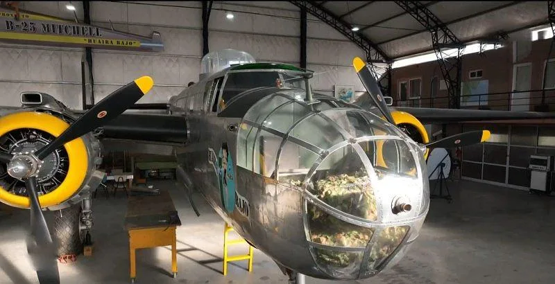 A front-right view of a silver B-25 Mitchell bomber with yellow propeller hubs, parked in a hangar.