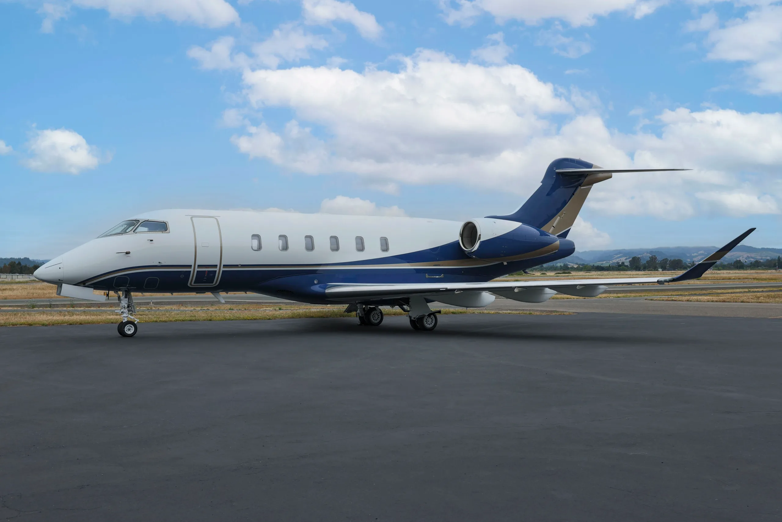 A side view of a Bombardier Challenger 350 business jet with white and blue livery parked on an airport tarmac.